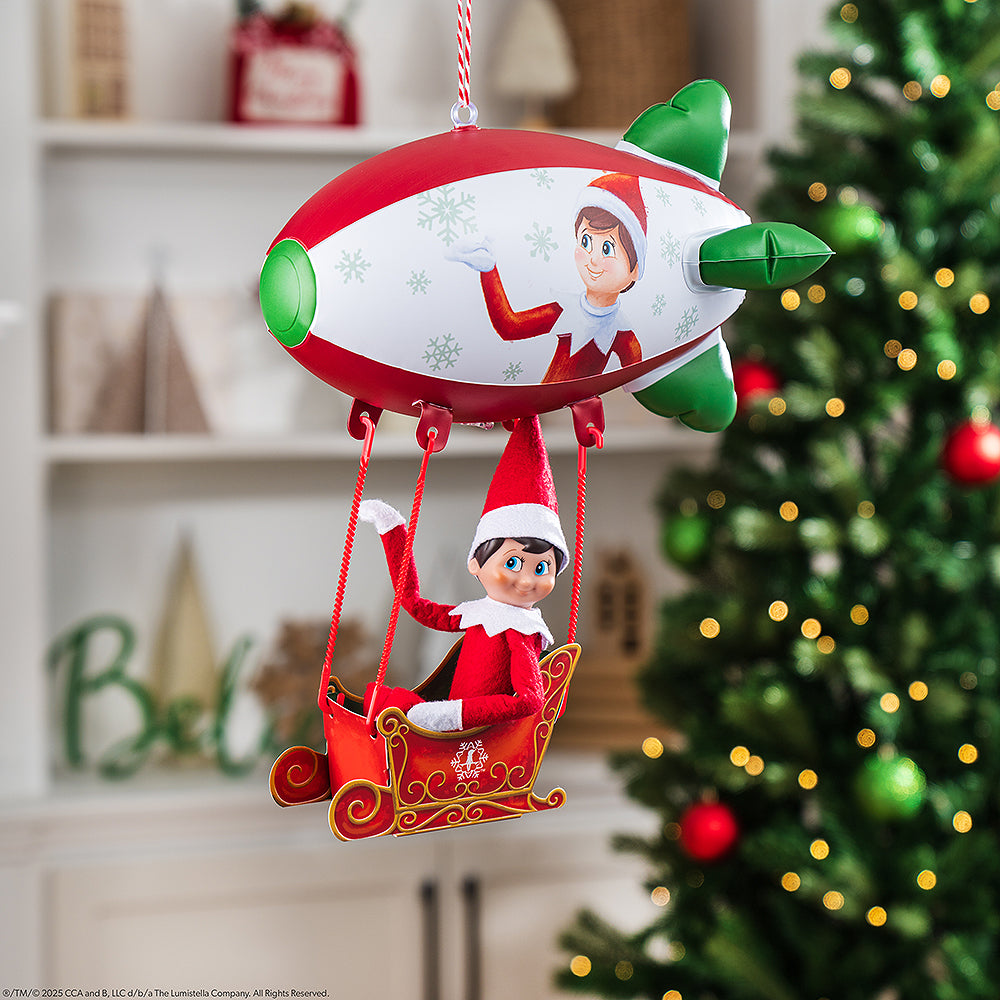 A boy Scout Elf® takes a ride and waves from the Polar Props™ Peppermint Blimp, with a Christmas tree and decor blurred in the background of this lifestyle image.