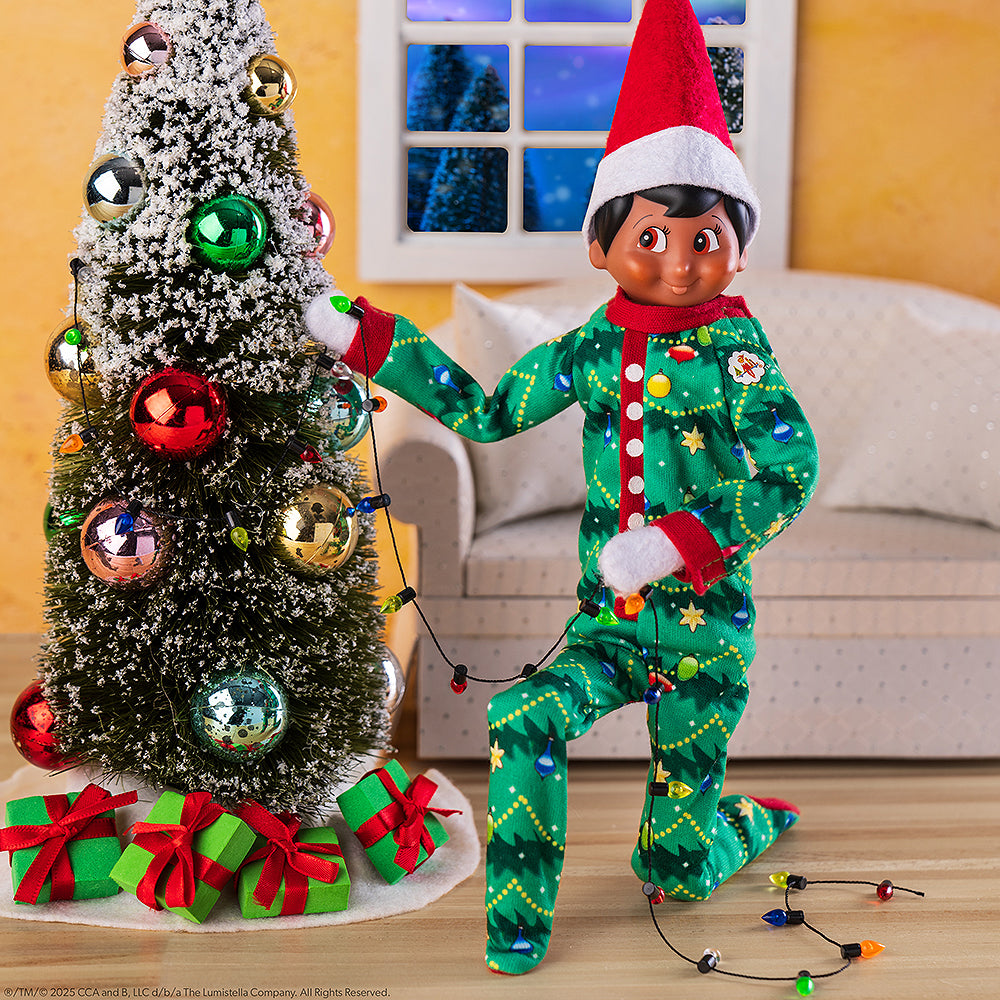 A scene of a boy Scout Elf donning the Claus Couture Collection® Goodnight, Sleep Bright PJs while wrapping Christmas lights on a miniature Christmas tree with ornaments and a little sofa in the background