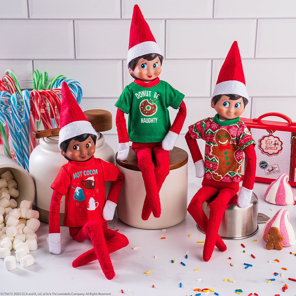 A trio of Scout Elves, each sporting a t-shirt from the Claus Couture Collection® Festive Fun T-Shirt Set, staged on a kitchen countertop surrounded by sweet, festive treats.