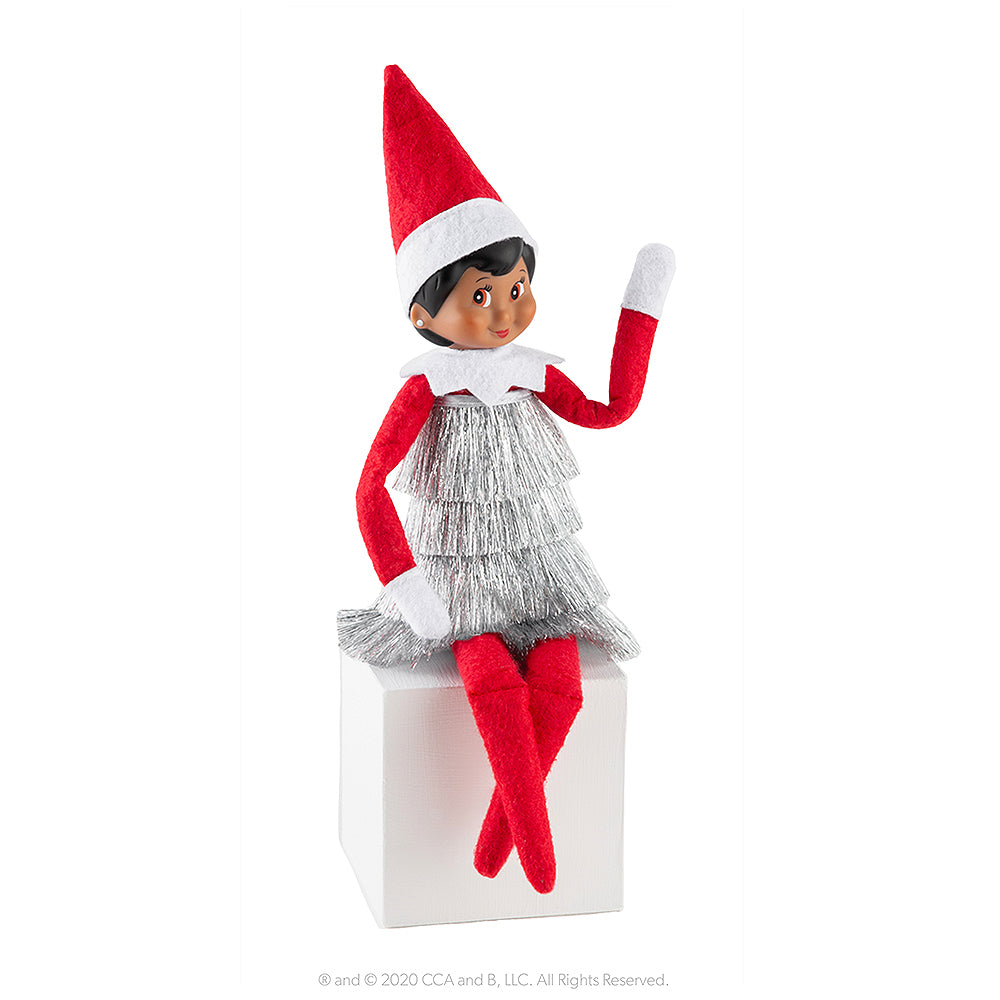 A girl Scout Elf® sits while modeling the Claus Couture Collection® Tiny Tinsel Dress while waving.