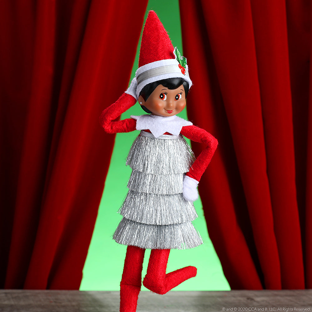 A girl Scout Elf® sports the Claus Couture Collection® Tiny Tinsel Dress in front of a red stage curtain in this lifestyle image.