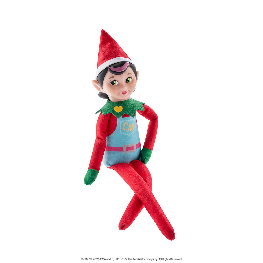 The Elf Mates® Chef girl doll poses sitting with her legs crossed and her left hand on her left hip. The Elf Mates Chef wears a red suit with green mittens and collar under a light blue apron with pink belt. The apron has a pocket with the EM insignia in yellow. Also in yellow is a heart on her green collar.