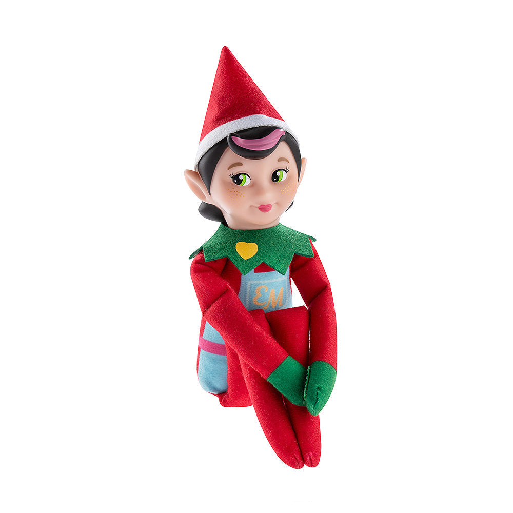 The Elf Mates® Chef girl doll poses sitting with her legs folded and her arms resting on her knees. The Elf Mates Chef wears a red suit with green mittens and collar under a light blue apron with pink belt. The apron has a pocket with the EM insignia in yellow. Also in yellow is a heart on her green collar.