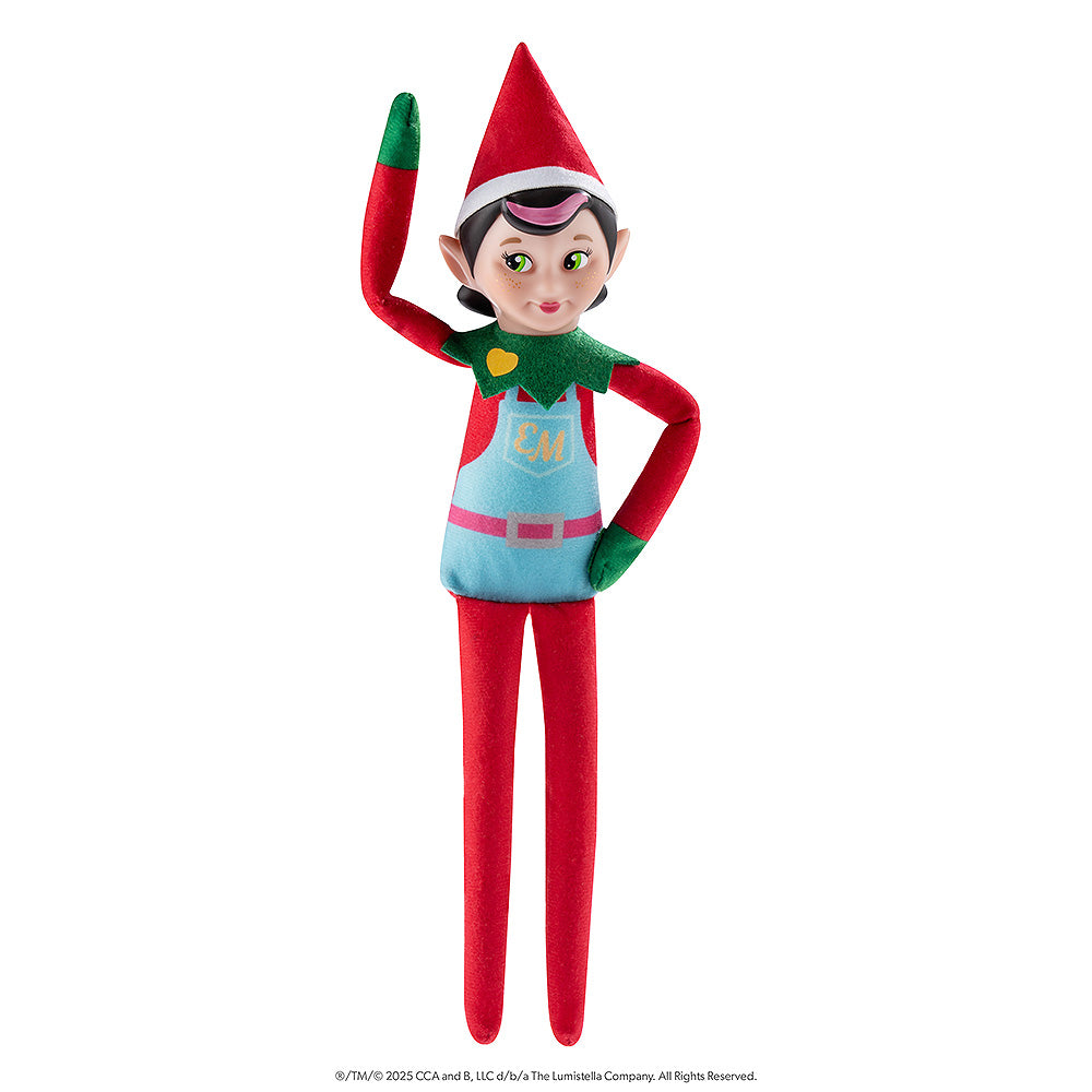 The Elf Mates® Chef girl doll poses standing while waving with her right hand and her left hand is resting on her hip. The Elf Mates Chef wears a red suit with green mittens and collar under a light blue apron with pink belt. The apron has a pocket with the EM insignia in yellow. Also in yellow is a heart on her green collar.