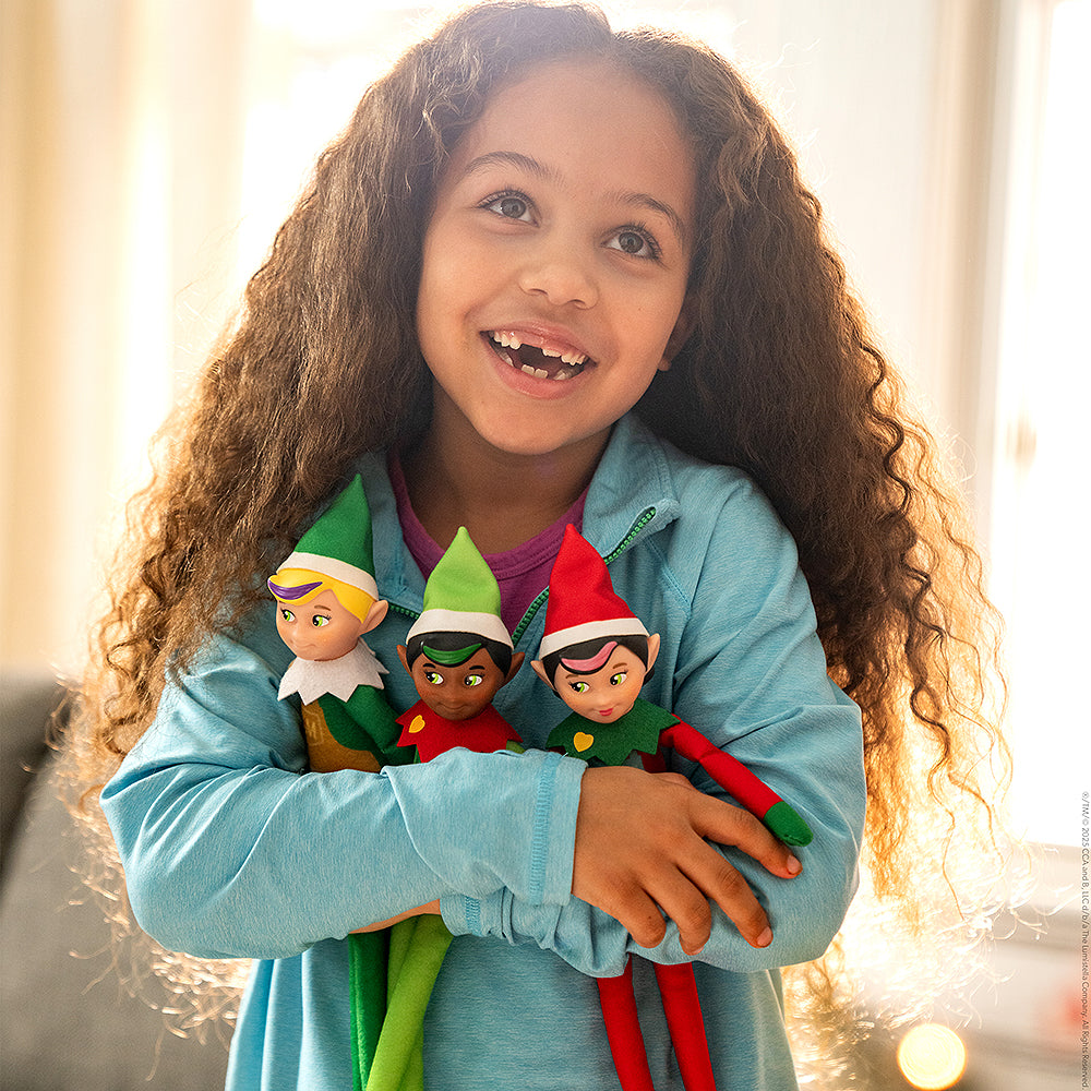 A young girl is clutching a trio of Elf Mates® dolls, including the Chef girl doll. She also has the Elf Mates Cobbler with blond hair and Toy Maker with black hair. She is standing in front of a window with golden light showering into the room, illuminating her. She is looking slightly upward to the left and smiling, revealing her missing teeth.