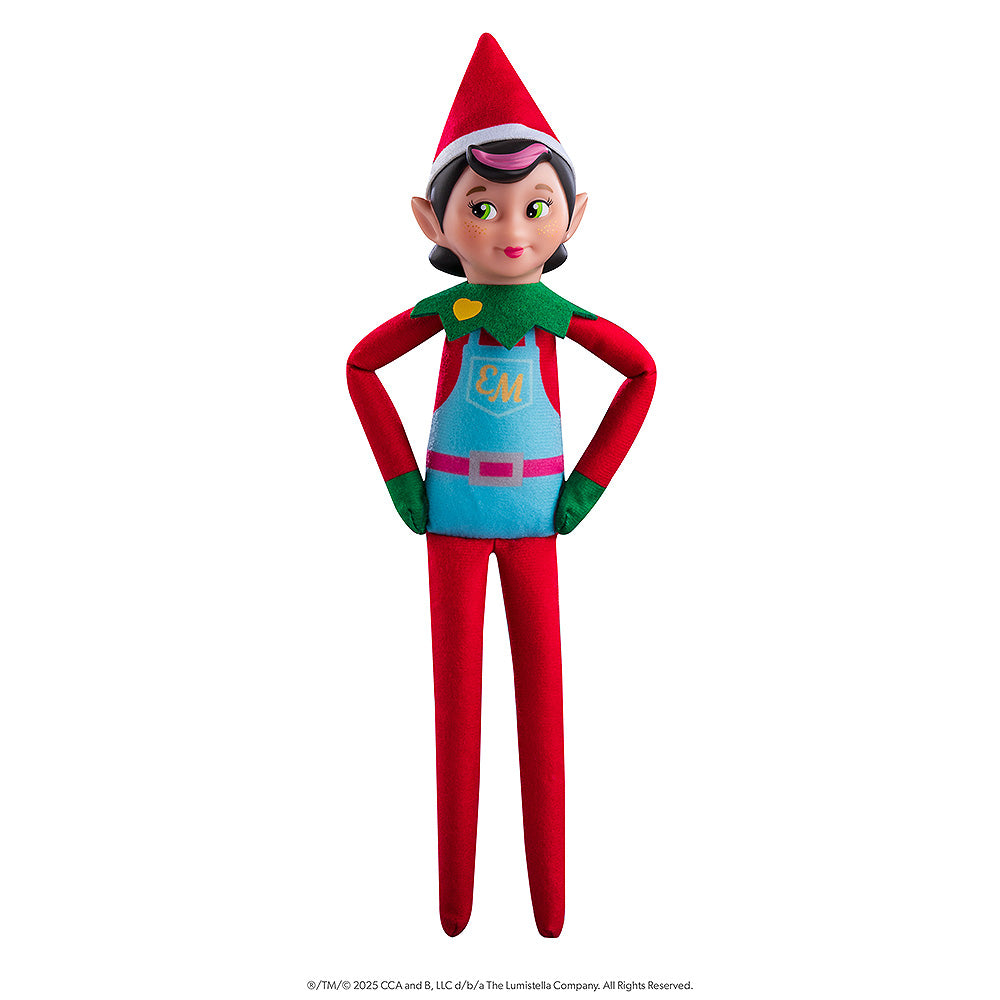 The Elf Mates® Chef girl doll poses standing while resting her hands on her hips. The Elf Mates Chef wears a red suit with green mittens and collar under a light blue apron with pink belt. The apron has a pocket with the EM insignia in yellow. Also in yellow is a heart on her green collar.