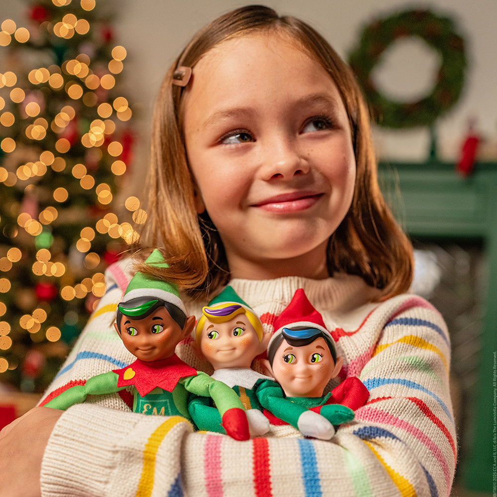 A young girl clutches a trio of Elf Mates® Cobbler, Toy Maker and Chef. She is wearing an off-white sweater with colored stripes. The background is blurred, but you can make out a lit, decorated Christmas tree, fireplace mantle, a wreath hanging overhead and a Scout Elf® sitting atop the mantle.