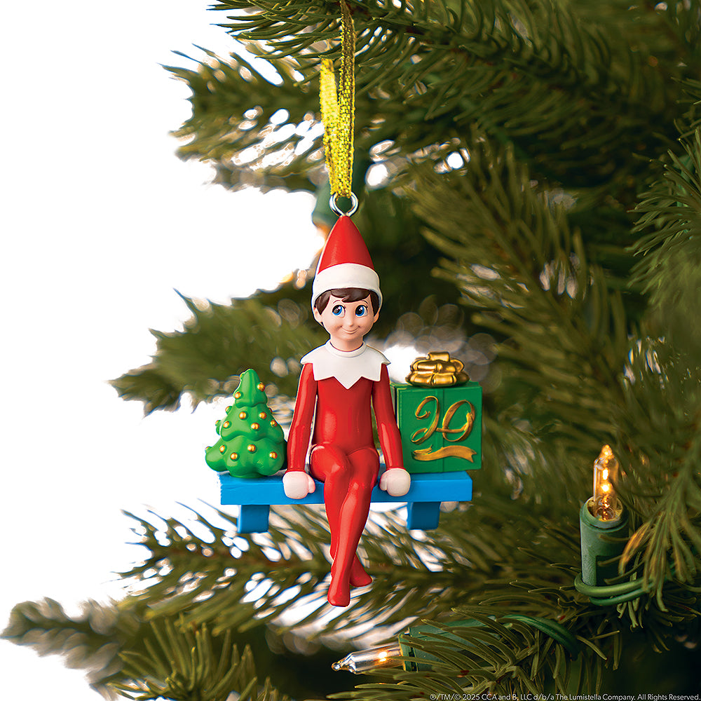 An up-close image of the included The Elf on the Shelf®: A Christmas Tradition, Limited 20th Anniversary Edition ornament hanging from a Christmas tree branch, featuring a Scout Elf sitting on the iconic blue shelf with a miniature Christmas tree to his right and a present wrapped in green paper to his left. The present has the 20th anniversary logo on it, adorned in a gold finish.
