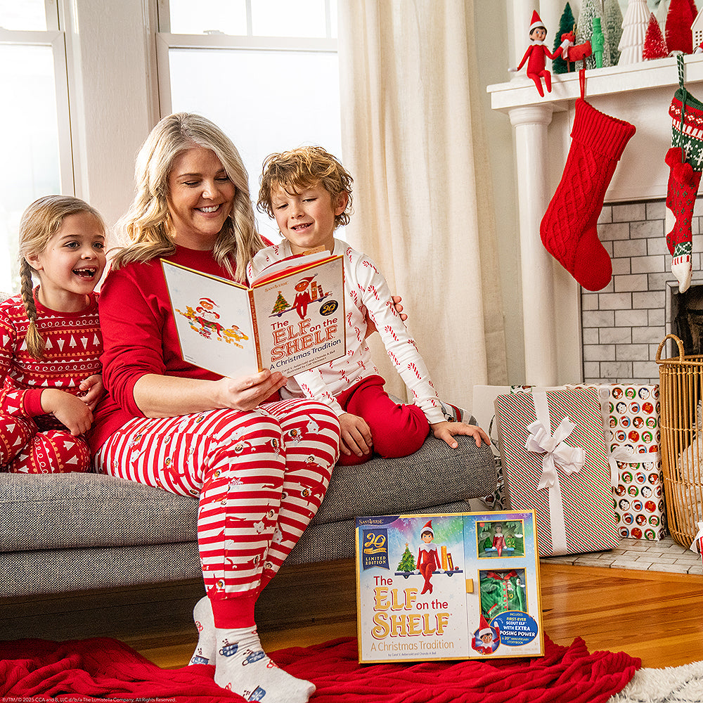 A lifestyle image of a mom and her daughter and son reading the The Elf on the Shelf®: A Christmas Tradition, Limited 20th Anniversary Edition book in their living room. They are seated on a sofa, dressed in Christmas pajamas, surrounded by vibrant Christmas decor. A Scout Elf sits on the mantle among Christmas tree decor and hanging stockings. Presents are in the foreground.