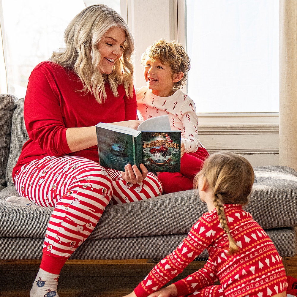 A mother and her two kids are huddled together in their living room reading The Rise of Nicholas the Noble. Her son is seated to her left, while her daughter is seated on the floor in front of her, facing her direction. They are being showered by light filtering in from the windows behind them. They are donning festive, Christmas PJs with red and white patterns with festive shapes and objects, even including a Scout Elf®.