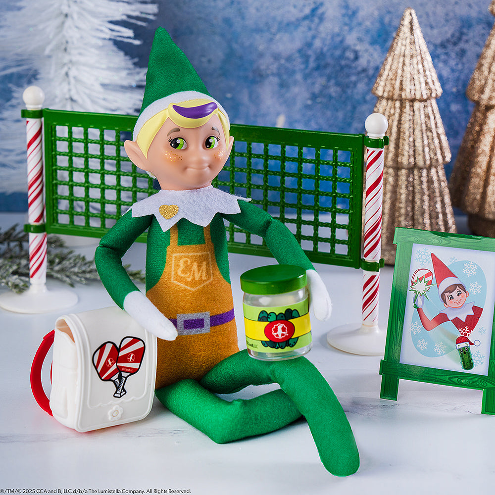 An Elf Mate Toy Maker gets in on the pickleball fun! He is staged with the included pack, pickle jar and sign, with the net and a festive background with trees behind him.