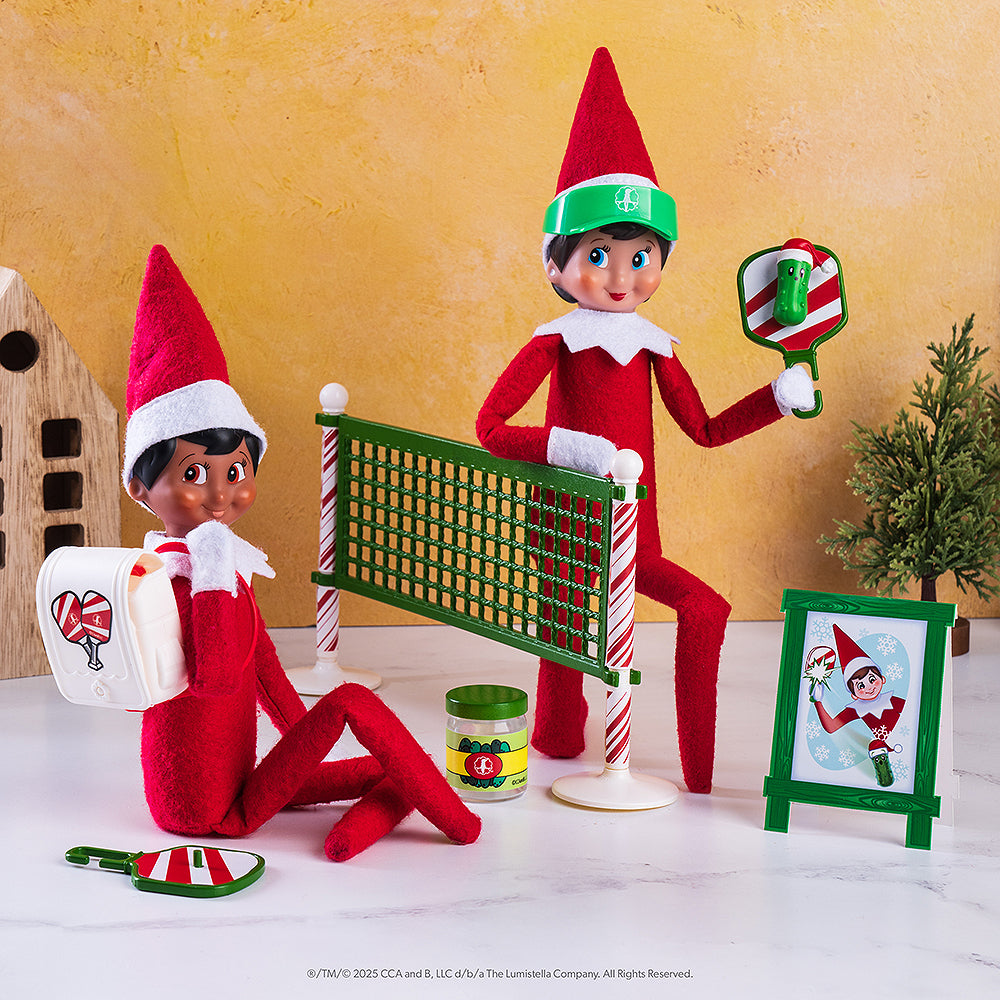A pair of Scout Elves posing with the Polar Props™ Pickleball Play Pack. A boy Scout Elf is sitting with the included pack strapped to his back, while a paddle is next to him on the ground near the the pickle jar. A girl Scout Elf is taking a knee by the net, holding up her paddle with the festive pickle attached to the racquet and the included pickleball sign to her right.