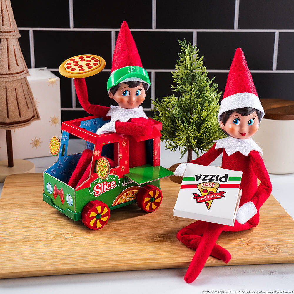A lifestyle image showing a pair of Scout Elves posing on a kitchen countertop and cutting board with the Polar Props™ Polar Pizza Truck set. There are miniature evergreen trees in the background.