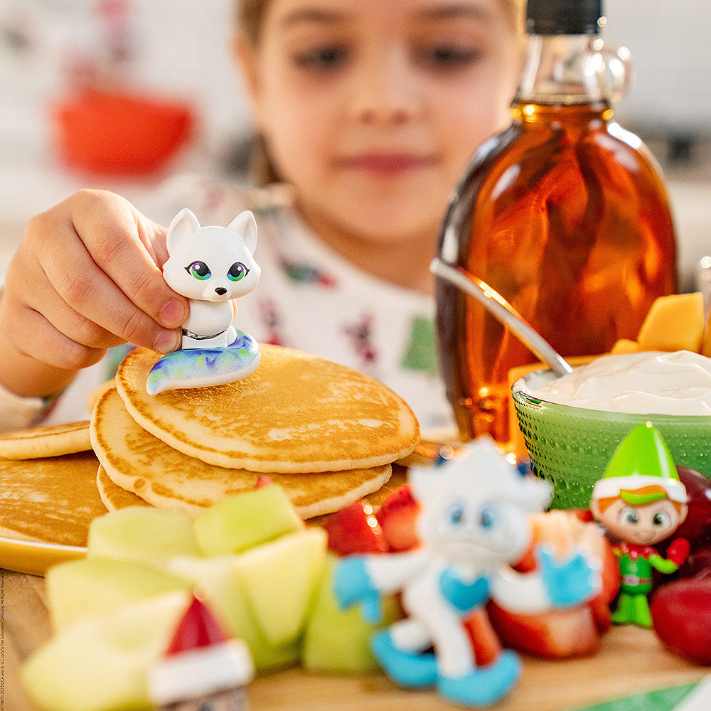 A young girl plays with the Santaverse™ Minis (Series 5) on a stack of pancakes at the kitchen table with fruit, syrup and other kitchen items shown.