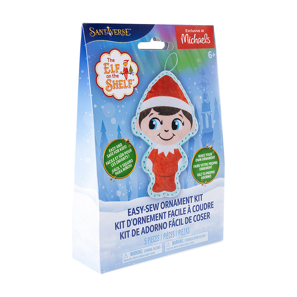 A front view of the packaging of The Elf on the Shelf® Easy-Sew Ornament Kit, which is slightly turned to the right. The packaging has the finished product, The Elf on the Shelf and Santaverse logos and other details. The packaging also has an "Exclusive at Michaels" label on it in the top right-hand corner. The background is a vector background commonly associated with the Santaverse brand, featuring conifer trees, light bursts, snowflakes, the Northern Lights, and a snow-covered landscape.