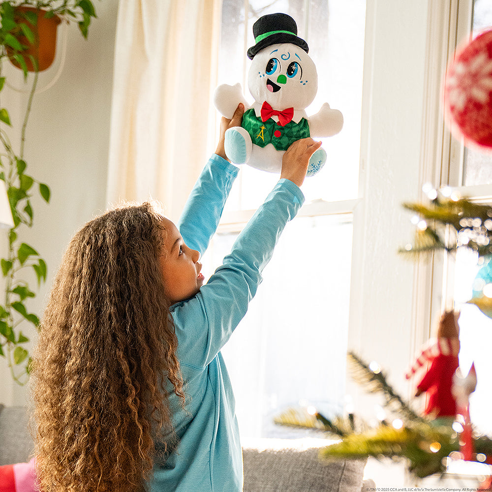 Lifestyle image of a little girl playing with SnoStax™ Razzle the Celebration Snowman plush in her home, decorated for Christmas