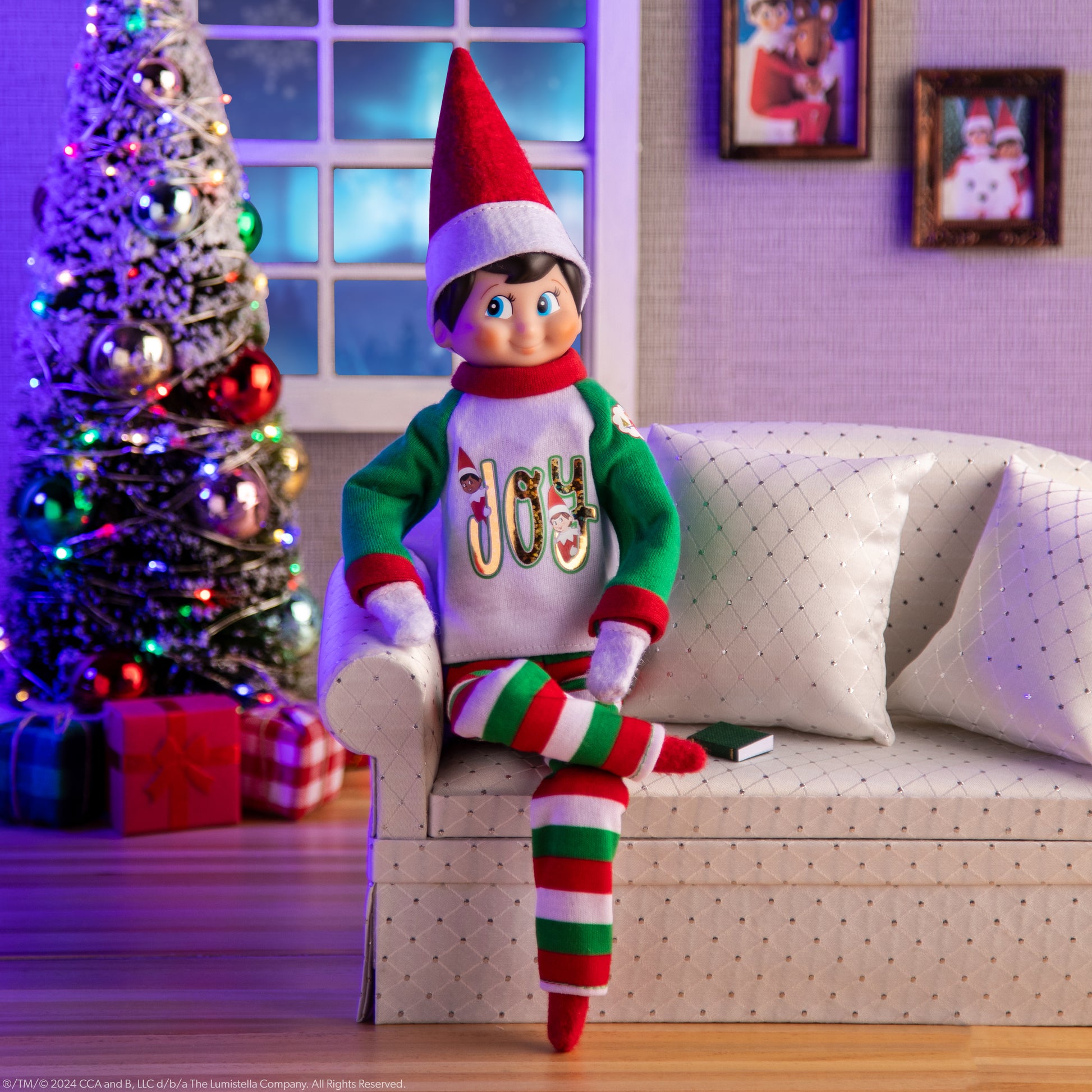 Scout Elf® sitting on the sofa wearing the Claus Couture Collection® Comfort & Joy PJs with a Christmas Tree and snowy window in the background 