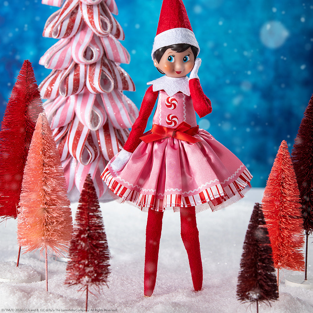 A girl Scout Elf® wearing the Claus Couture Collection® Pink Peppermint Party Dress staged in a snowy forest with a candy cane color palette