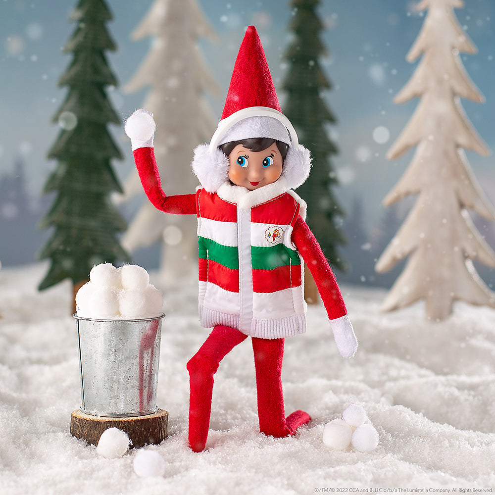 A girl Scout Elf® wearing the Claus Couture Collection® Bundle-Up Puffer Vest and earmuffs, staged in a snowy forest with a galvanized bucket filled with snowballs. She is cocking her arm back to throw a snowball in a friendly snowball fight.