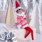 A girl Scout Elf is seated on a wrapped Christmas gift wearing the Claus Couture Collection® Snowflake Sweetie Dress. She is surrounded by matching Christmas decor of little miniature conifer trees that appear to be wrapped in coordinating wrapping paper patterns and a tinsel background and silver bows.