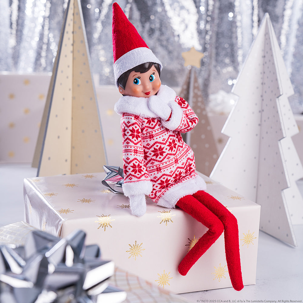 A girl Scout Elf is seated on a wrapped Christmas gift wearing the Claus Couture Collection® Snowflake Sweetie Dress. She is surrounded by matching Christmas decor of little miniature conifer trees that appear to be wrapped in coordinating wrapping paper patterns and a tinsel background and silver bows.