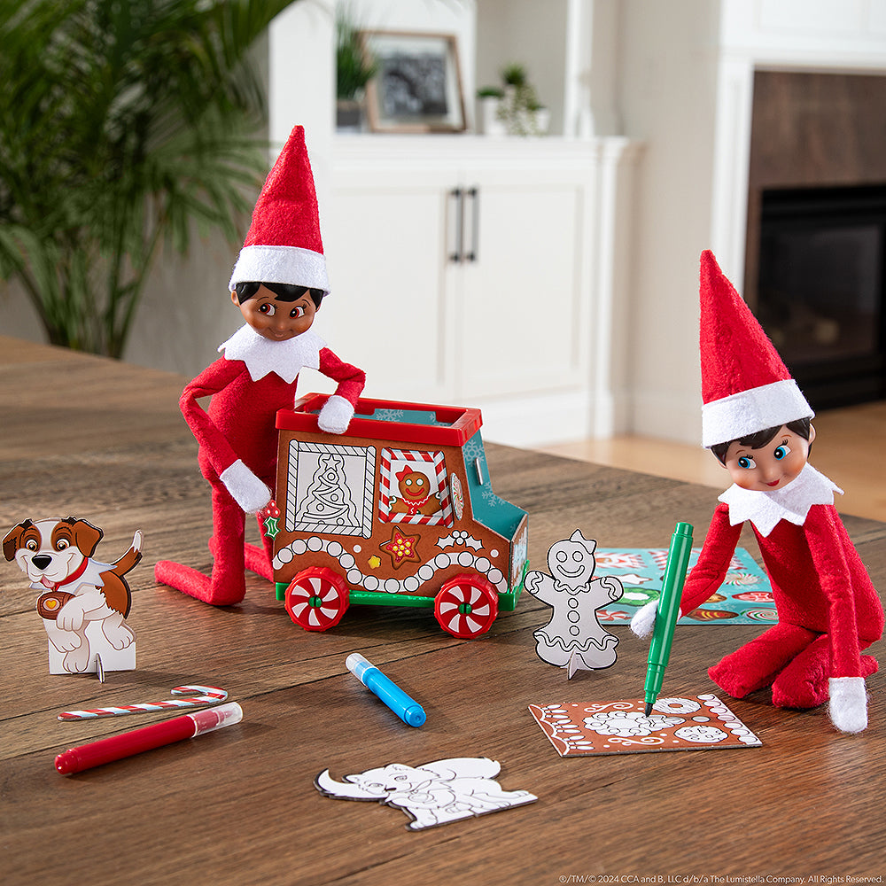 A pair of Scout Elves® staged next to the truck and other props, one of the Scout Elves® is using the marker to color in a gingerbread panel that makes the truck