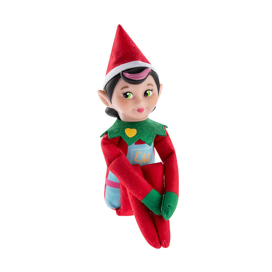 The Elf Mates® Chef girl doll poses sitting with her legs folded and her arms resting on her knees. The Elf Mates Chef wears a red suit with green mittens and collar under a light blue apron with pink belt. The apron has a pocket with the EM insignia in yellow. Also in yellow is a heart on her green collar.