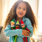 A young girl is clutching a trio of Elf Mates® dolls, including the Chef girl doll. She also has the Elf Mates Cobbler  with blond hair and Toy Maker with black hair. She is standing in front of a window with golden light showering into the room, illuminating her. She is looking slightly upward to the left and smiling, revealing her missing teeth.
