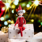 A lifestyle image of the included Scout Elf® boy with blue eyes. He is posed sitting on a pile of wrapped presets, wrapped in white wrapping paper with gold flourishes and a metallic gold bow. In the background, you can see the glimmer of lights on a Christmas tree. Most notably, the Scout Elf is adorned from head to toe in Swarovski® crystals that coordinate with his signature style.