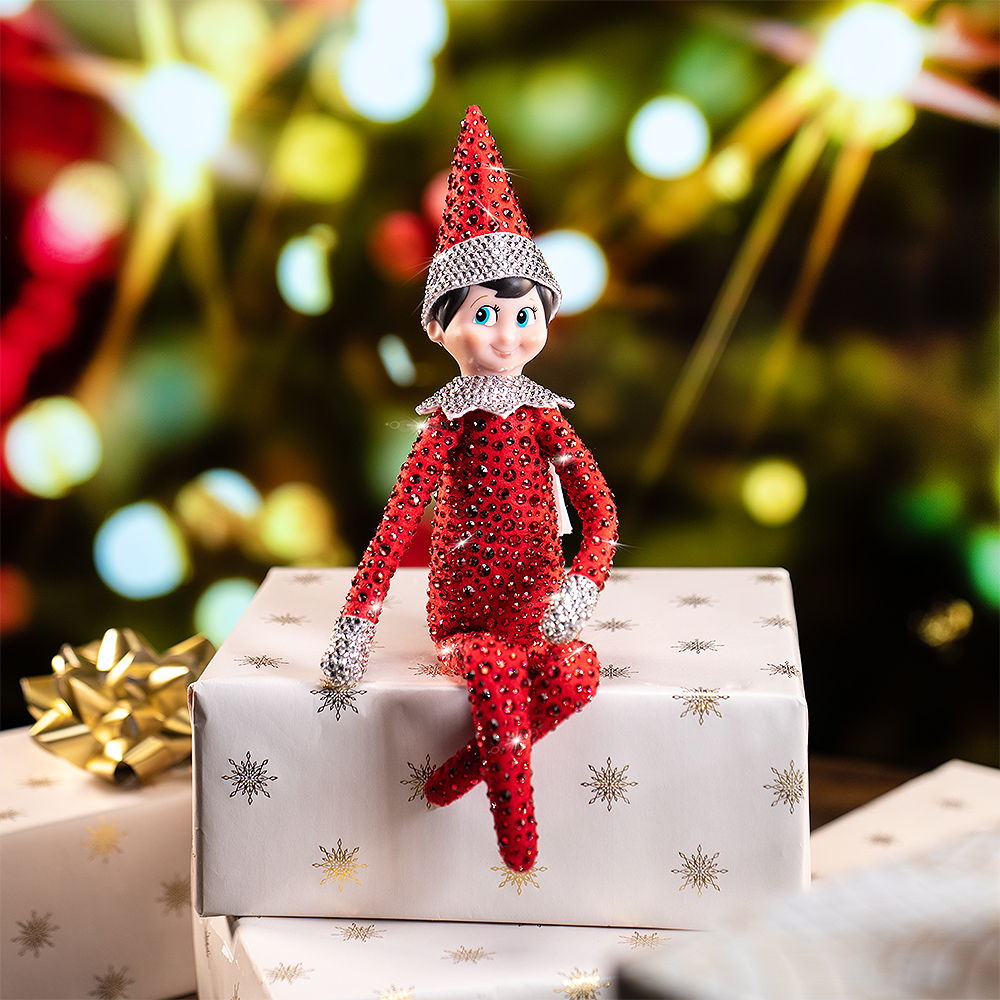 A lifestyle image of the included Scout Elf® boy with blue eyes. He is posed sitting on a pile of wrapped presets, wrapped in white wrapping paper with gold flourishes and a metallic gold bow. In the background, you can see the glimmer of lights on a Christmas tree. Most notably, the Scout Elf is adorned from head to toe in Swarovski® crystals that coordinate with his signature style.