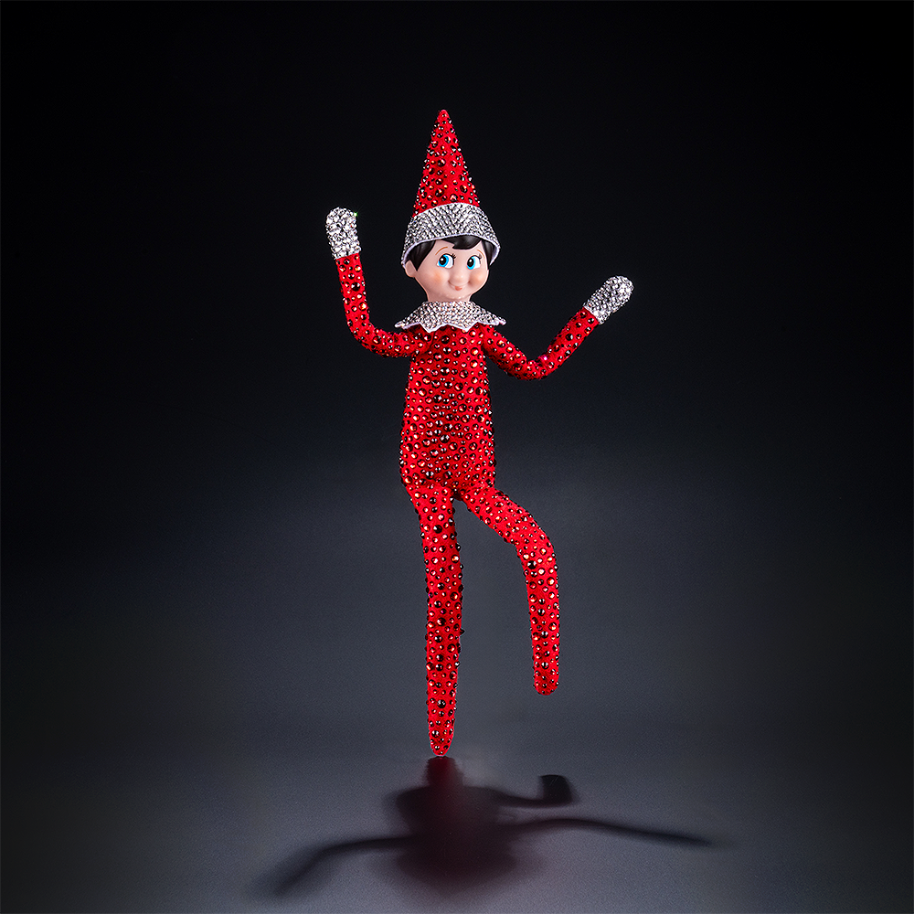 The included Scout Elf® boy with blue eyes poses to show off his flexible bending limbs for even easier elf scenes. Most notably, he is adorned from head to toe in Swarovski® crystals that coordinate with his signature style. A dark background surrounds him, with a spotlight shining down on his figure.