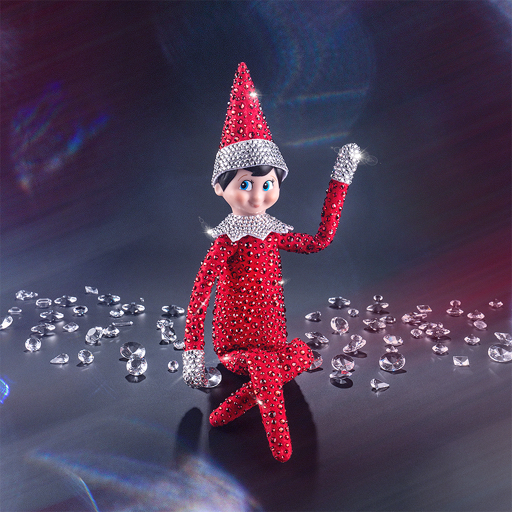 A lifestyle shot of the included Scout Elf® boy with blue eyes poses to show off his flexible bending limbs for even easier elf scenes. Most notably, he is adorned from head to toe in Swarovski® crystals that coordinate with his signature style. A dark background with refracted light surrounds him, which shows up with red and blue hues.