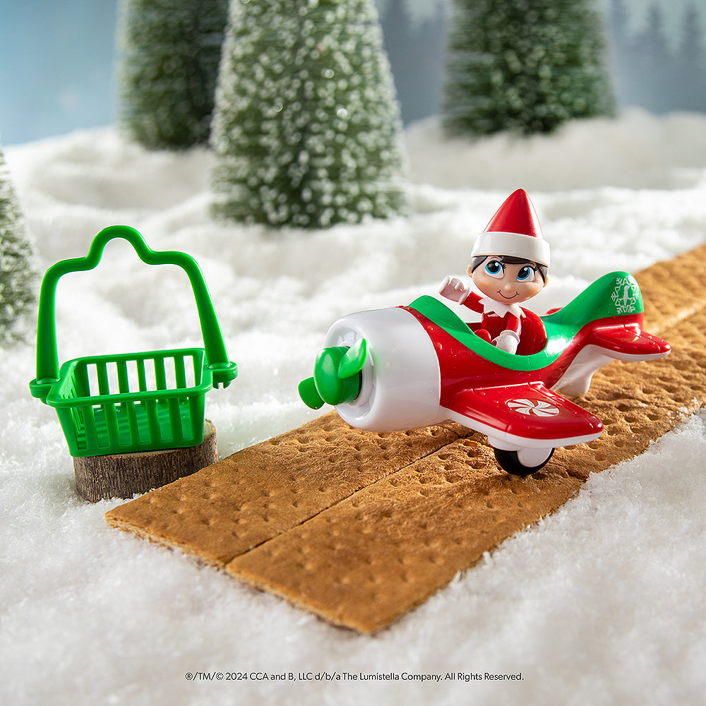 The Elf on the Shelf® Action Figure Play Pack: Flight Edition items staged in a snowy forest with graham crackers acting as a runway for the plane