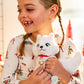 A little girl adorned in the Elf on the Shelf pajamas is snuggling her Arctic Fox plush. Christmas decor can be seen in the background, but it is obscured.