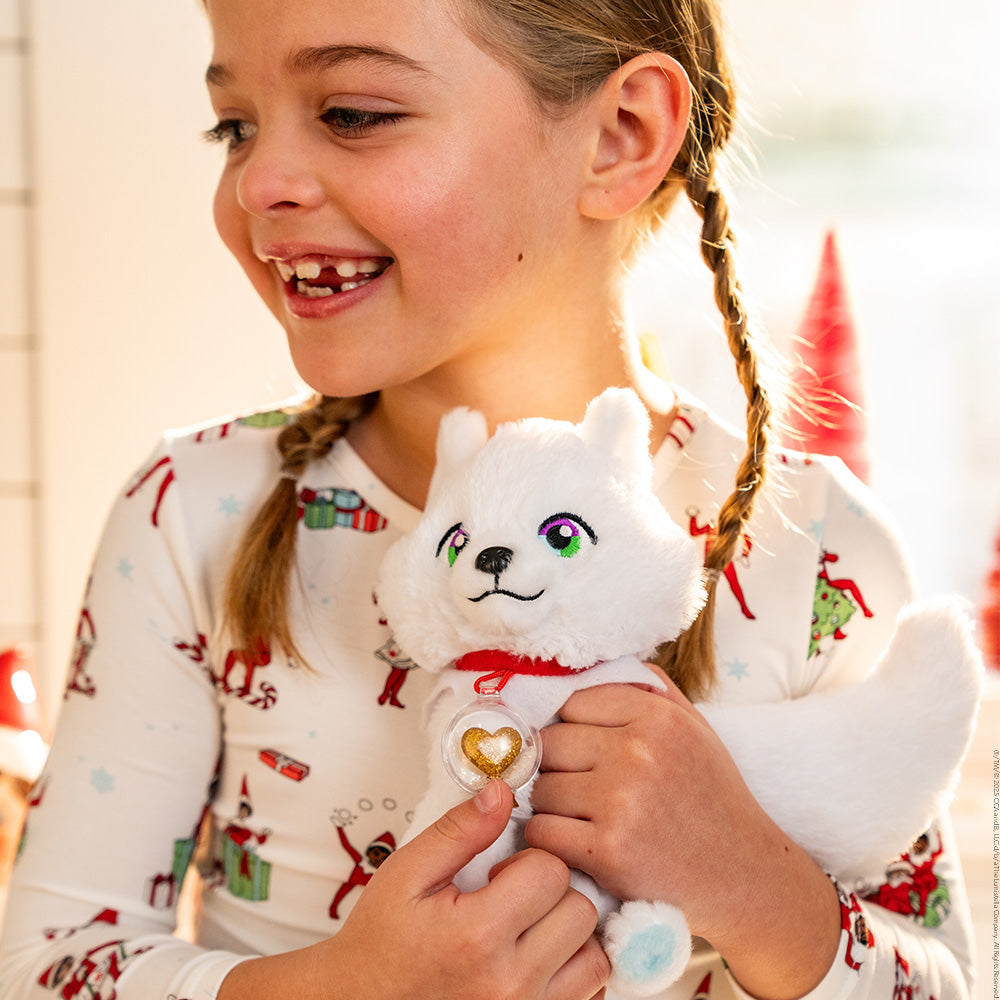 A little girl adorned in the Elf on the Shelf pajamas is snuggling her Arctic Fox plush. Christmas decor can be seen in the background, but it is obscured.