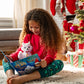 A little girl reads her Elf Pets®: An Arctic Fox Tradition storybook with her Arctic Fox plush sitting in her lap. She is seated on the living room rug in her home and you can see the Christmas decor, tree and stockings hanging in the background. A Scout Elf can also be spotted in the background.
