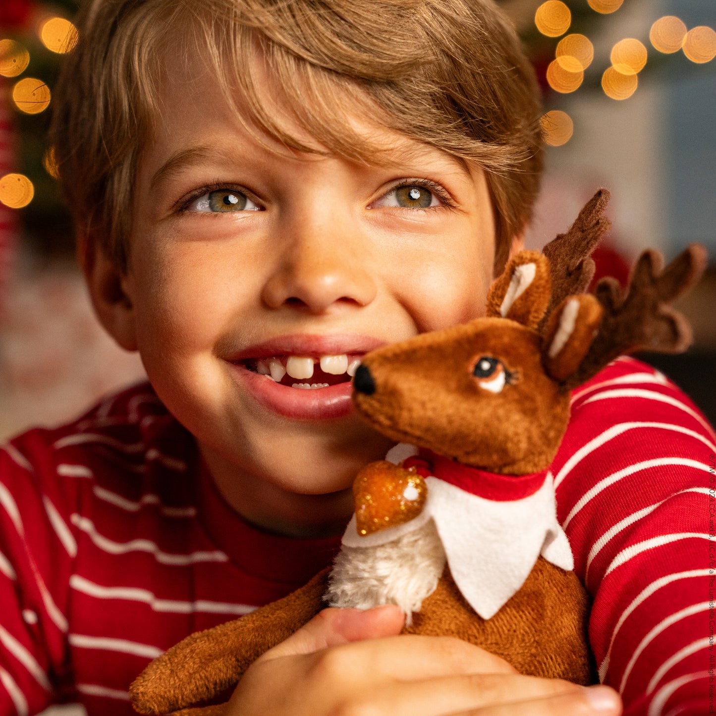 A little boy snuggles his Elf Pets Reindeer with Christmas lights out of focus in the background