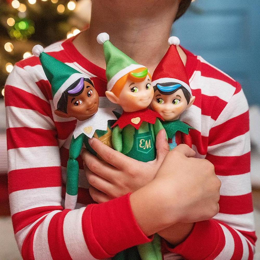 A little boy in Christmas pajamas snuggling the Elf Mates® Chef, Cobbler and Toy Maker dolls with a Christmas tree in the background