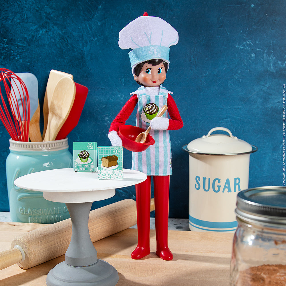 A staged Scout Elf® baking up yummy treats while sporting the MagiFreez® Christmas Chef outfit amongst a rolling pin and other baking tools and supplies