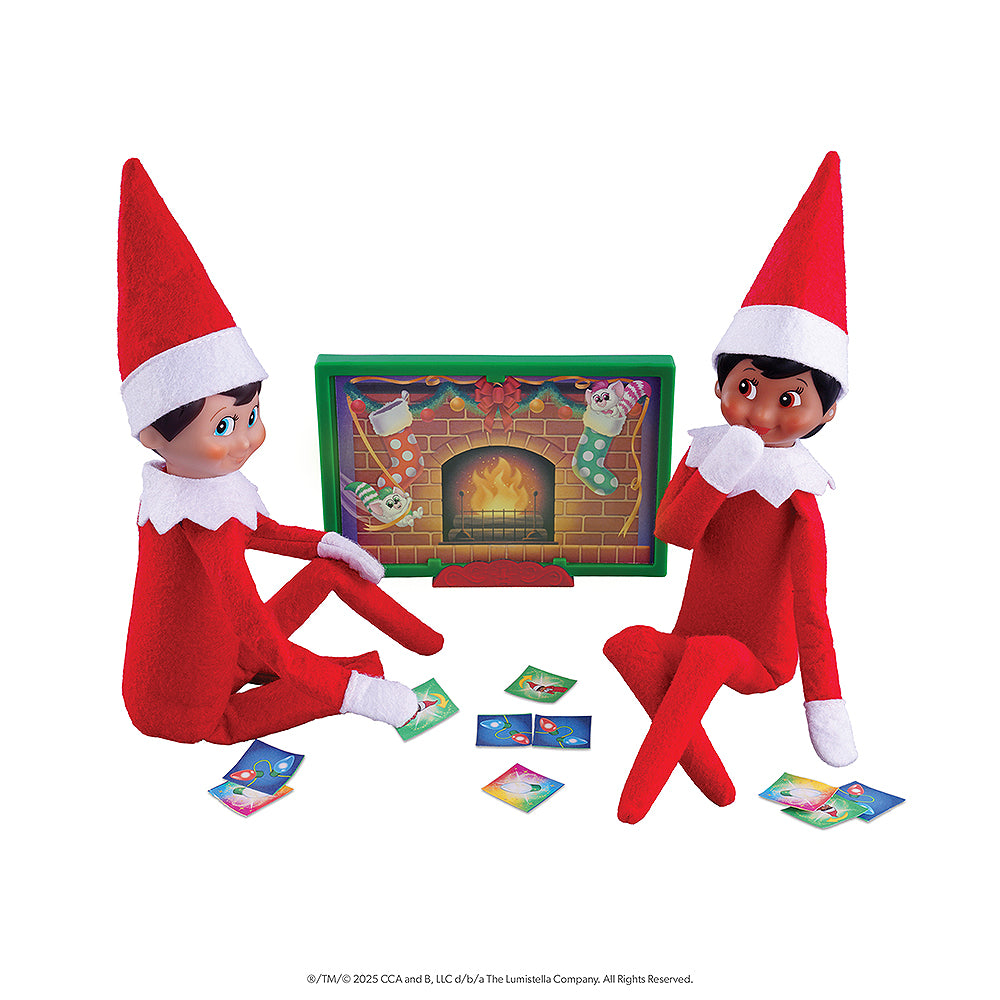 A pair of Scout Elves are posed in front of a fireplace prop while playing a card game, all of which are included in the Polar Props™ The Official Elf Ideas Fun & Games Kit.