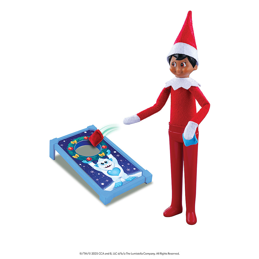 A boy Scout Elf is playing with a festive corn hole board, featuring the one and only SnoBiggie Yeti Lil Grunt on the board. The Scout Elf is posed tossing a red bean bag and holding a blue bean bag in his other hand. All of the items shown are included in the Polar Props™ The Official Elf Ideas Fun & Games Kit.