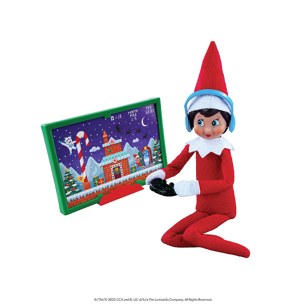 A girl Scout Elf is posing with a game controller and earphones, with a TV screen showing a fun and festive game. The details on the screen show a wintery setting with a SnoBiggie Yeti Lil Grunt and a Scout Elf. These items are included in the Polar Props™ The Official Elf Ideas Fun & Games Kit.