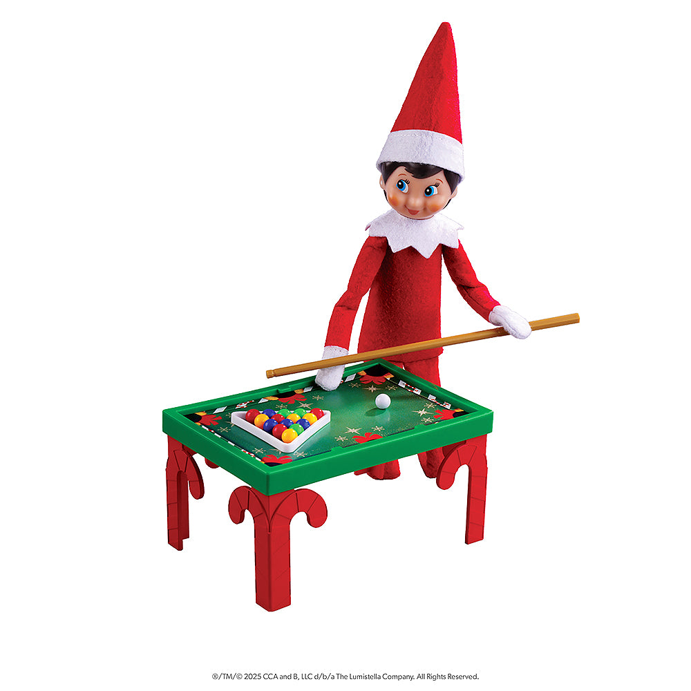 A girl Scout Elf is playing pool with a pool cue and pool balls on a festive looking pool table. These props are included in the Polar Props™ The Official Elf Ideas Fun & Games Kit.