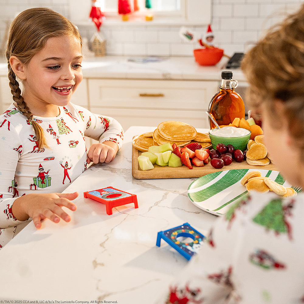 A lifestyle image showing a pair of siblings  wearing The Elf on the Shelf PJs, while playing one of the games from the Polar Props™ The Official Elf Ideas Fun & Games Kit. they are playing in their kitchen at the kitchen island, adjacent to a cutting board with breakfast foods consisting of pancakes, syrup, fruit and yogurt. In the background are a pair of Scout Elves staged on the kitchen counter.