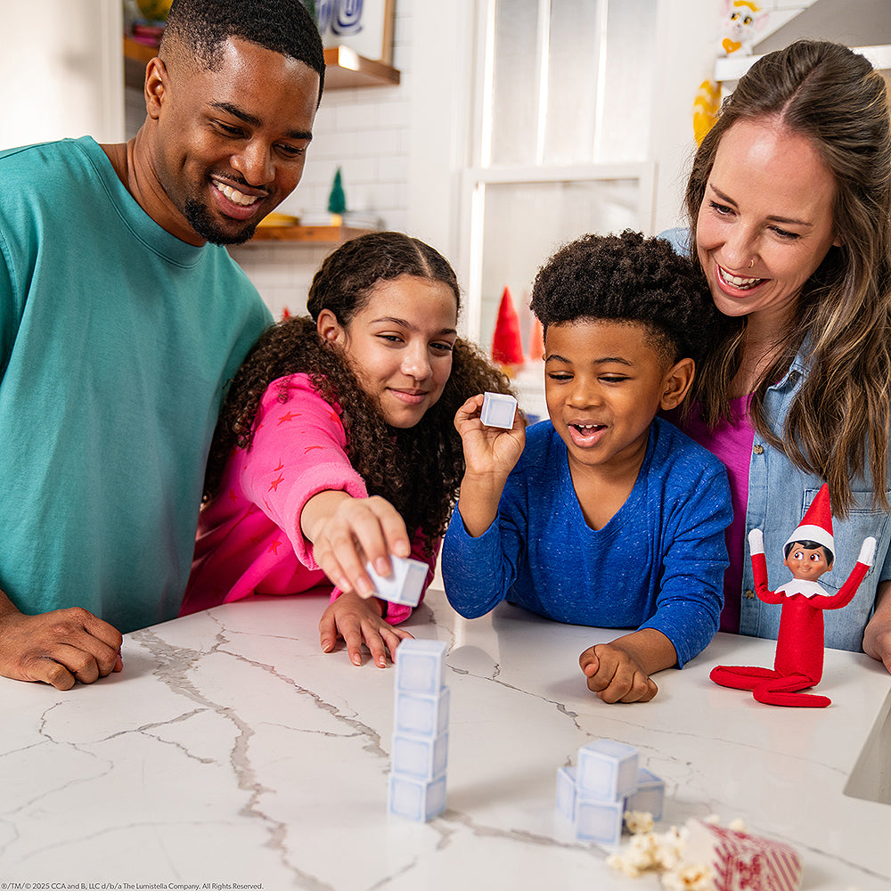 A lifestyle image of a family playing one of the games from the Polar Props™ The Official Elf Ideas Fun & Games Kit. They are gathered around a coffee table playing the game. A Scout Elf is also propped on the table watching them play with his arms posed in the air, as though he is one of the family members himself and is cheering on the children playing the game with their parents flanking them in the background.