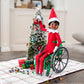 A Scout Elf® sitting in the Polar Props™ Elfcare Mobility Set wheelchair, decorating a mini Christmas tree