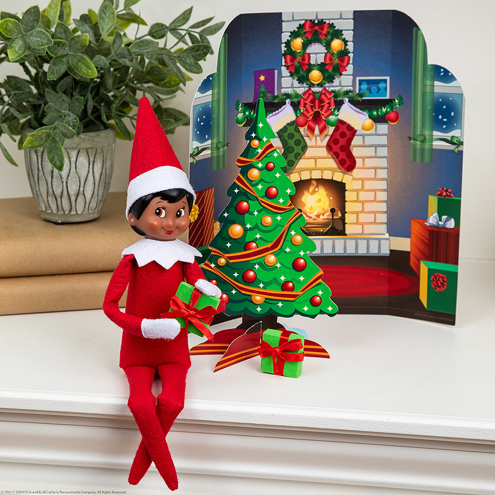 A girl Scout Elf® staged with the Polar Props™ 24-Day Ultimate Elf Ideas Kit items from Day 10, which is a fireplace backdrop, a Christmas tree, and 2 tiny presents