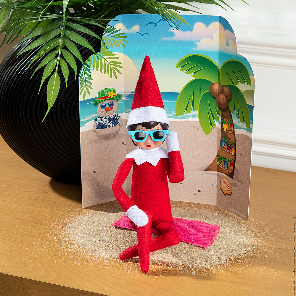 A girl Scout Elf® staged with the Polar Props™ 24-Day Ultimate Elf Ideas Kit items from Day 18, which is a tropical beach background with a snowman made out of sand, and a tiny beach towel that the Scout Elf® is sitting on with sunglasses