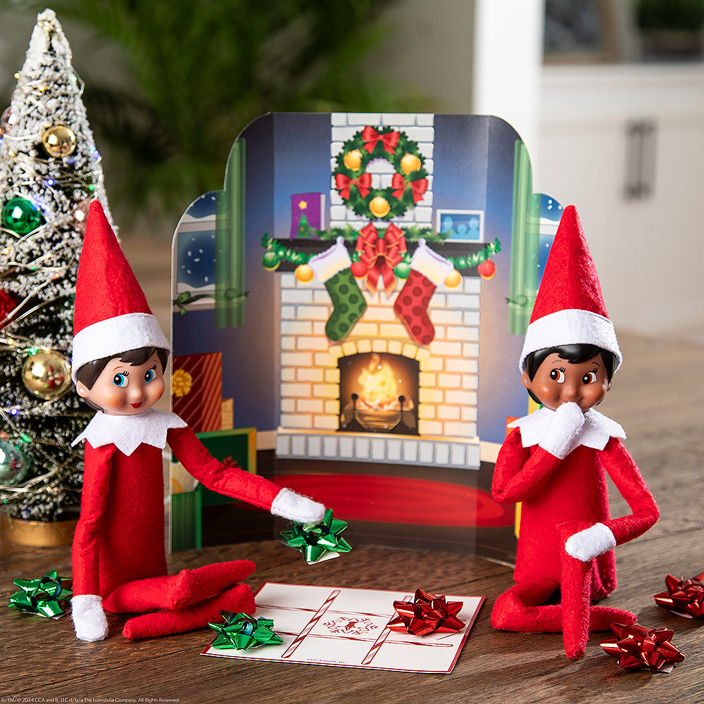 A pair or Scout Elves® staged with the Polar Props™ 24-Day Ultimate Elf Ideas Kit items from Day 7, which is a fireplace backdrop and a tic-tac-toe board that they're playing using tiny wrapping bows