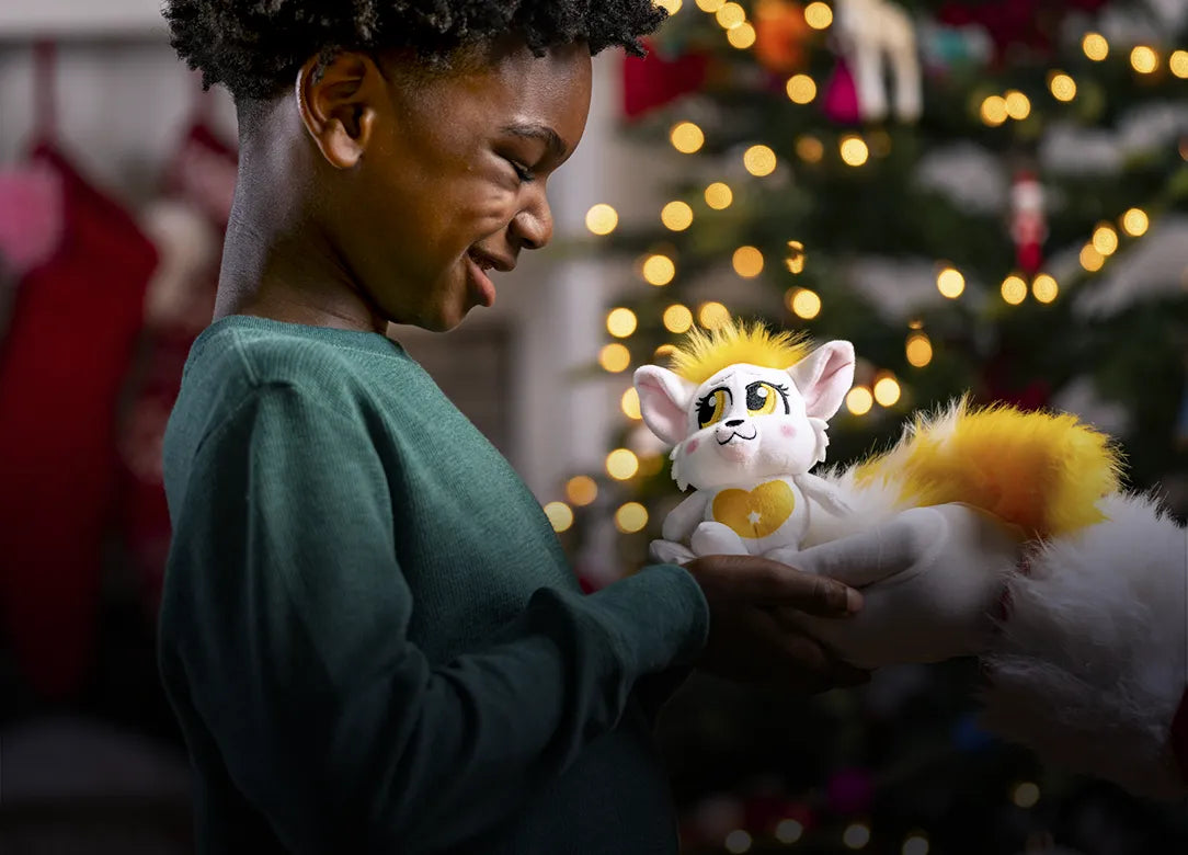 Hands of Santa Claus holding a Yellow Bogie and giving it to a smiling boy. In the background is a Christmas Tree, mantel, and stockings.